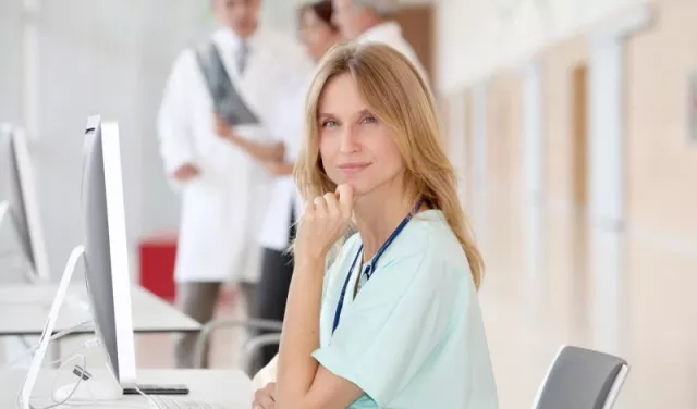 Doctor in baby blue shirt wearing stethoscope around neck, sitting in front of computer, looking into the camera.